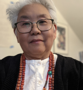 Mushim Ikeda wearing glasses, a white shirt, black cardigan and red beads