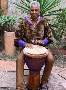 Image of a Black woman wearing a striped shirt and brown pants, seated with a hand drum.