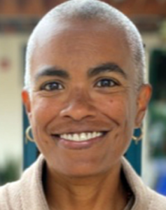 Photo of Tania Triana, a brown-skinned person with short gray hair, wearing circular earrings and smiling.
