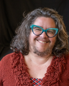 Image of Dawn Haney, a White person with teal glasses, shoulder-length brown hair, and facial hair, smiling and wearing a red top.