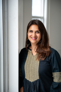 Photo of Anjali Sawhney, a South Asian woman with medium brown hair, wearing a green and beige dress and smiling