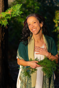 Photo of Renata Moreira, wearing a white dress with a dark green and beige shawl, holding a bunch of herbs and smiling