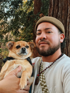 Photo of Andres Gonzalez, a mixed heritage person with facial hair, wearing a beanie, white shirt, beads, and holding a small brown dog