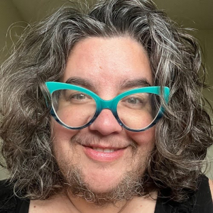 Dawn Haney, with shoulder length wavy grayish hair, turquoise glasses, and light facial hair